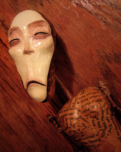 A paper mache doll made of antique book pages and sticks, with a long pale face, squinting sadly into the distance with a thin frown