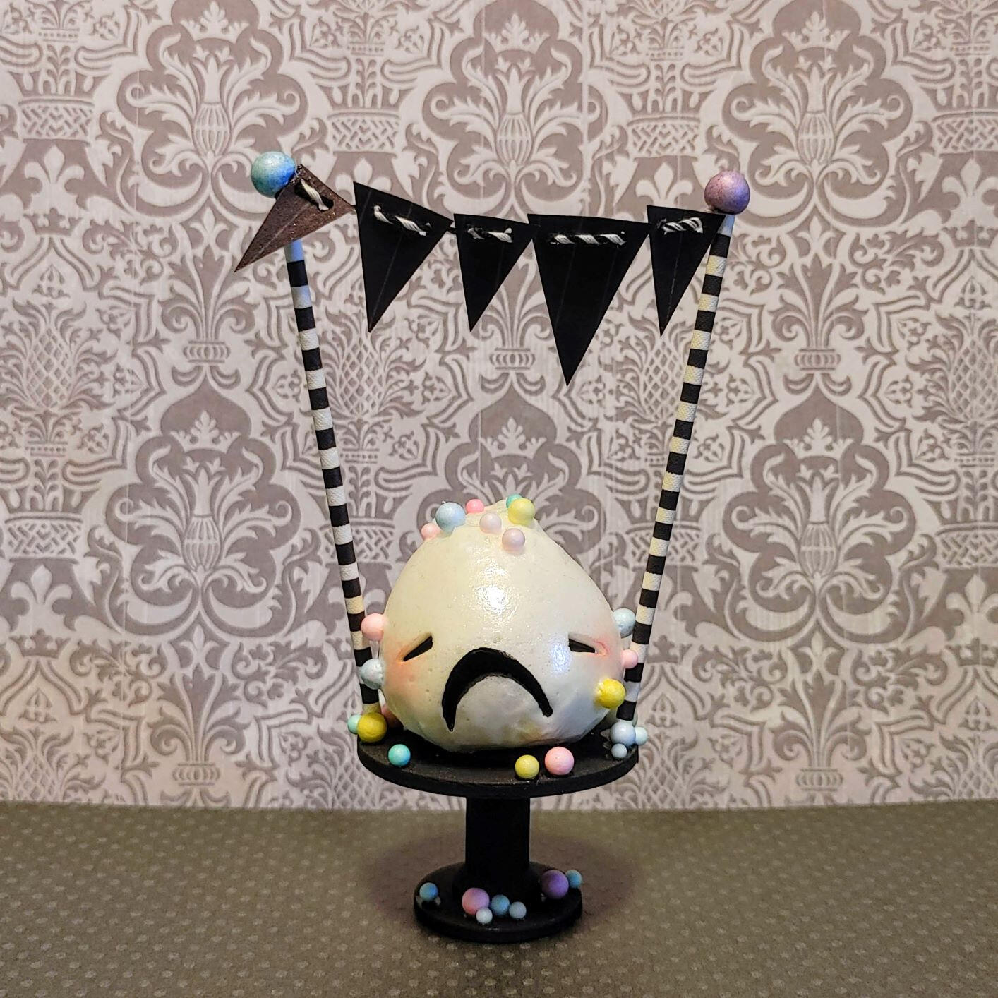 Small handmade sculpture of a wailing frosting creature covered in colorful sprinkles and displayed on a tiny cake pedestal.