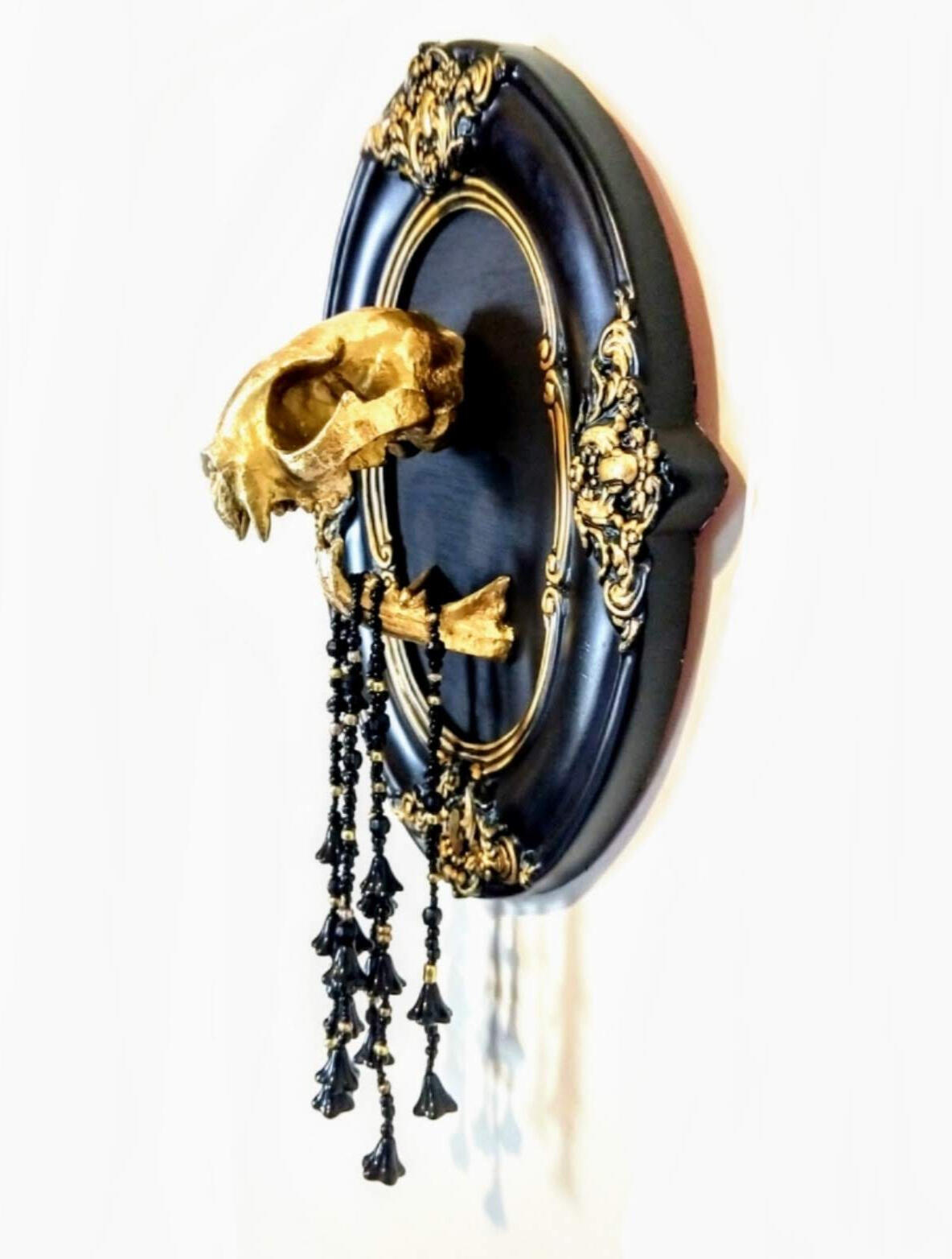 A gold (synthetic) cats skull mounted in an ornate black and gold oval victorian frame, with black beaded strands dripping with black glass flowers hanging from its jaws