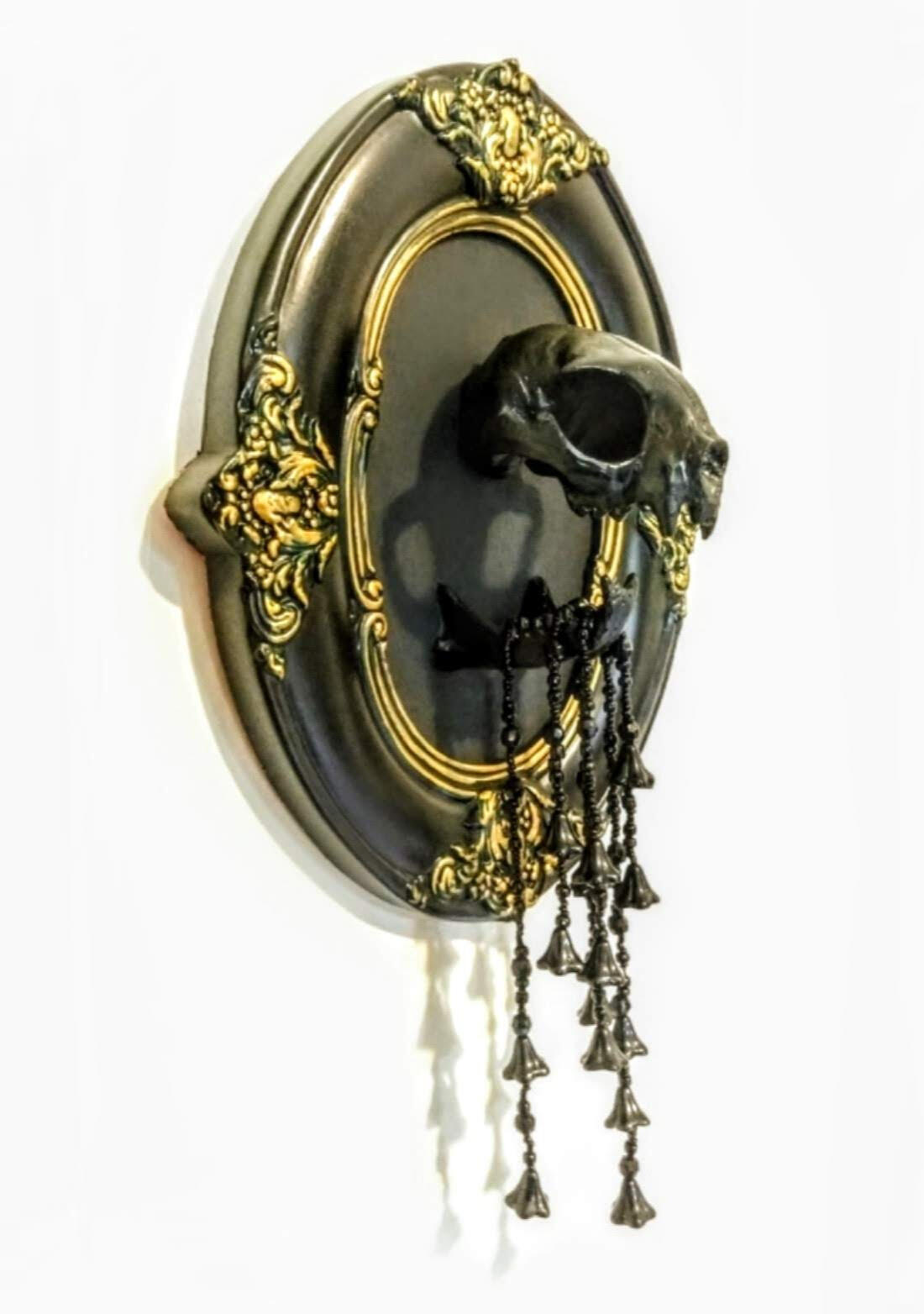 A black (synthetic) cats skull mounted in an ornate black and gold oval victorian frame, with black beaded strands dripping with black glass flowers hanging from its jaws