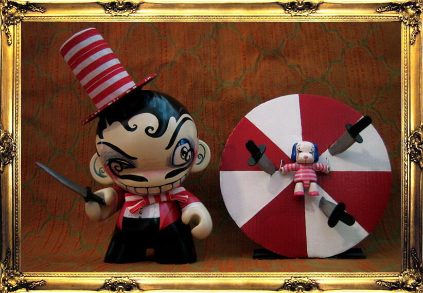 The Knife Thrower and His (First) Assistant A Munny and Dunny, painted to be circus performers - a grinning knife thrower wearing a tall red and white striped hat holding a knife and squinting sideways at their assistant - a small blue eared dog wearing a matching outfit, tied to a spinning red and